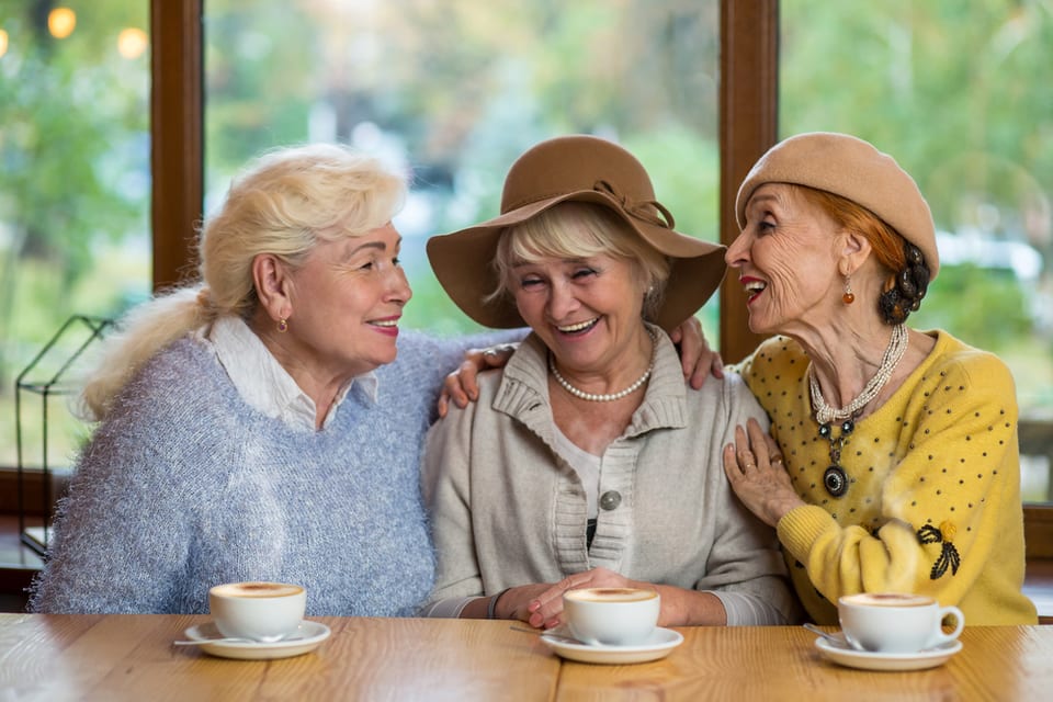 Three senior women laugh and talk while drinking coffee.