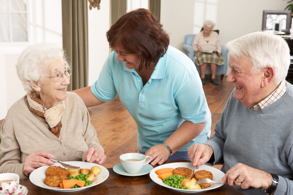 A senior man and woman smile while eating a nutritious meal at an assisted living facility.