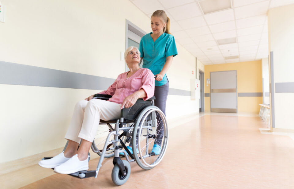 A caregiver helping a senior woman in a wheelchair through a facility hallway