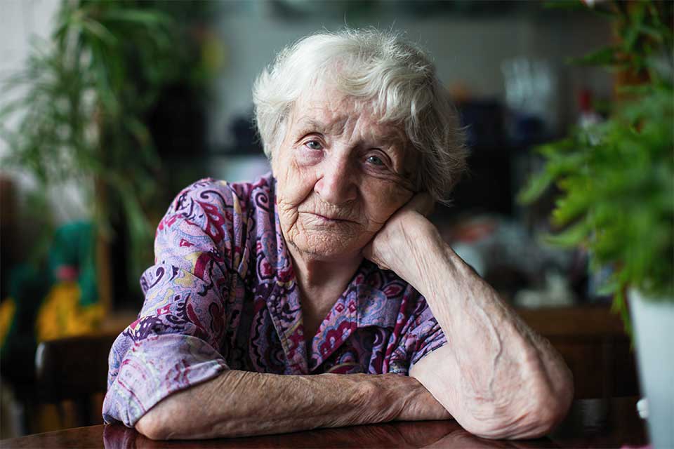 Elderly woman seated at a table resting her head on her hand.